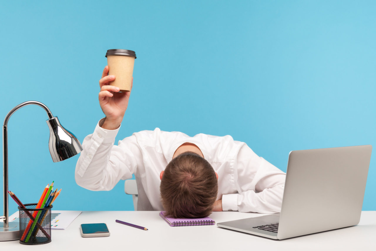 Man in white button up with head on desk holding up a coffee cup. Laptop, notebook, phone, pencils and a lamp are on the desk surrounding him with a plain blue background.