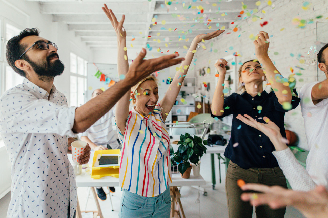 A group of coworkers throwing confetti in the air at the office, all with smiles on their faces.