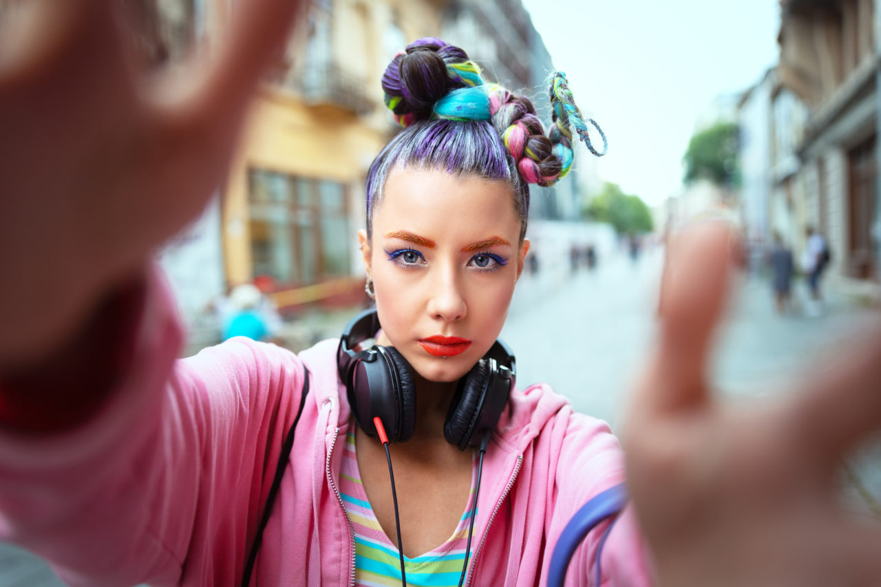 A young girl with colourful hair, bright makeup, pink jacket and headphones around her neck looking into the camera with hands out towards the camera