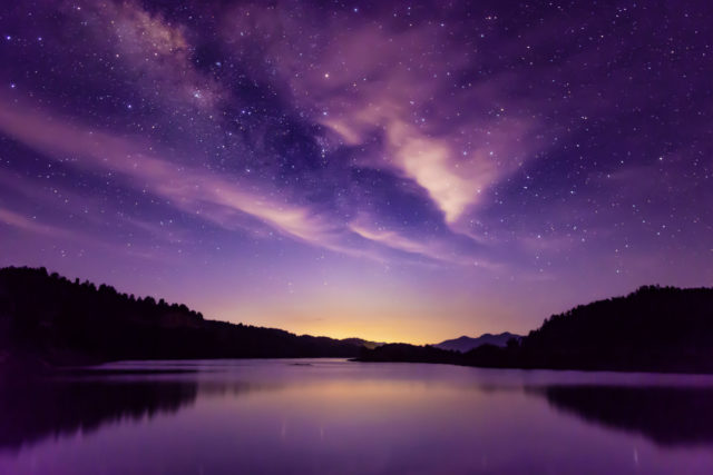 A purple lake with mountains in the background on the horizon blacked out from the purple milky way in the sky with lots of little stars