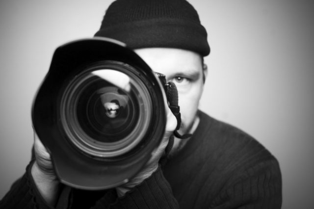 A black and white photo of a photographer holding a huge lens close to the camera, he is behind the camera with only one eye showing and is wearing a beanie.