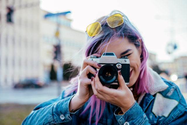 A young girl with purple hair taking a photo using an old camera pointing towards the camera, she has yellow sunglasses on her head and is wearing a denim jacket.
