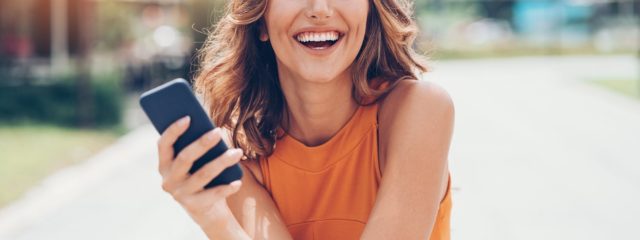 A young girl smiling wearing an orange tank top holding her phone