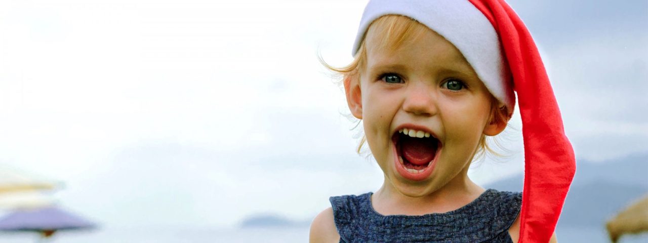 A little girl wearing a santa hat with her mouth open smiling at the camera