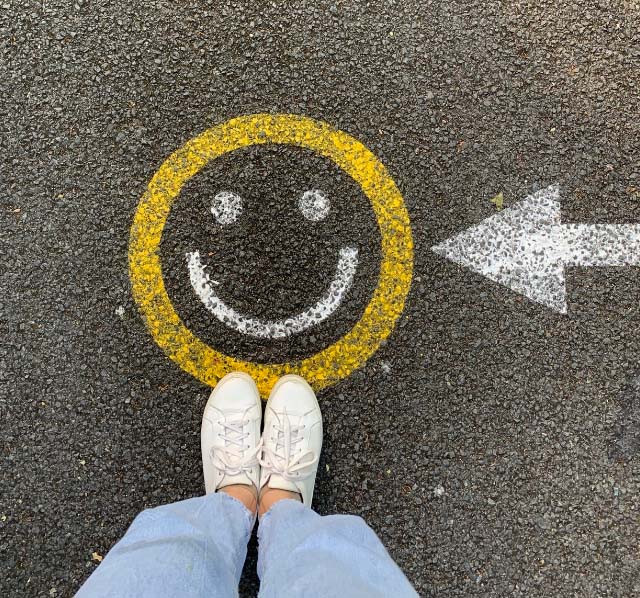 A smiley face with an arrow pointed at it spray painted onto the road with a girls shoes and jeans shown standing beside it.