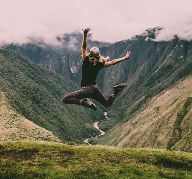A girl in athletic wear jumping in the air at the top of a mountain range, down below is a stream of water in between mountains.